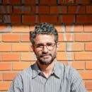 Outdoor portrait of a man with eyeglasses posing against a sunlit brick wall.